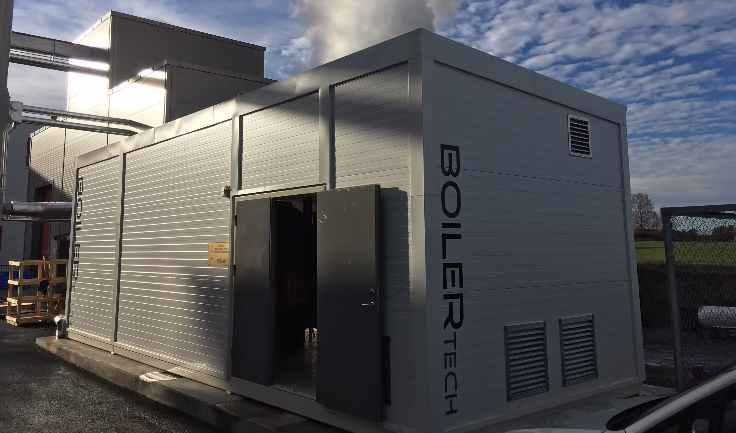 Modular boiler container from Boilertech with open doors, ventilation grilles, and a pipe connection to an industrial building.