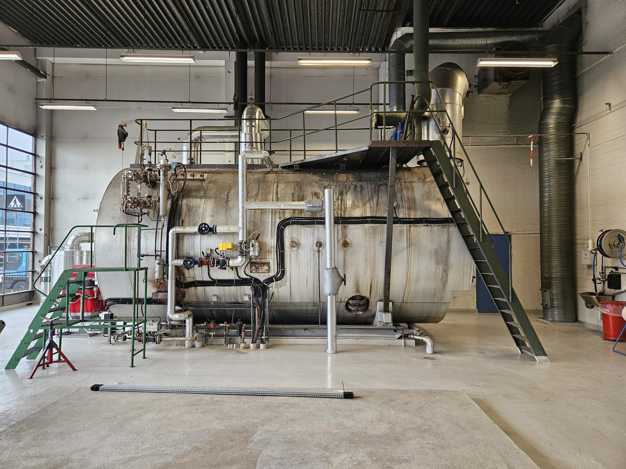 En stor industriell kjele med rør, ventiler og en trapp i et fabrikklokale