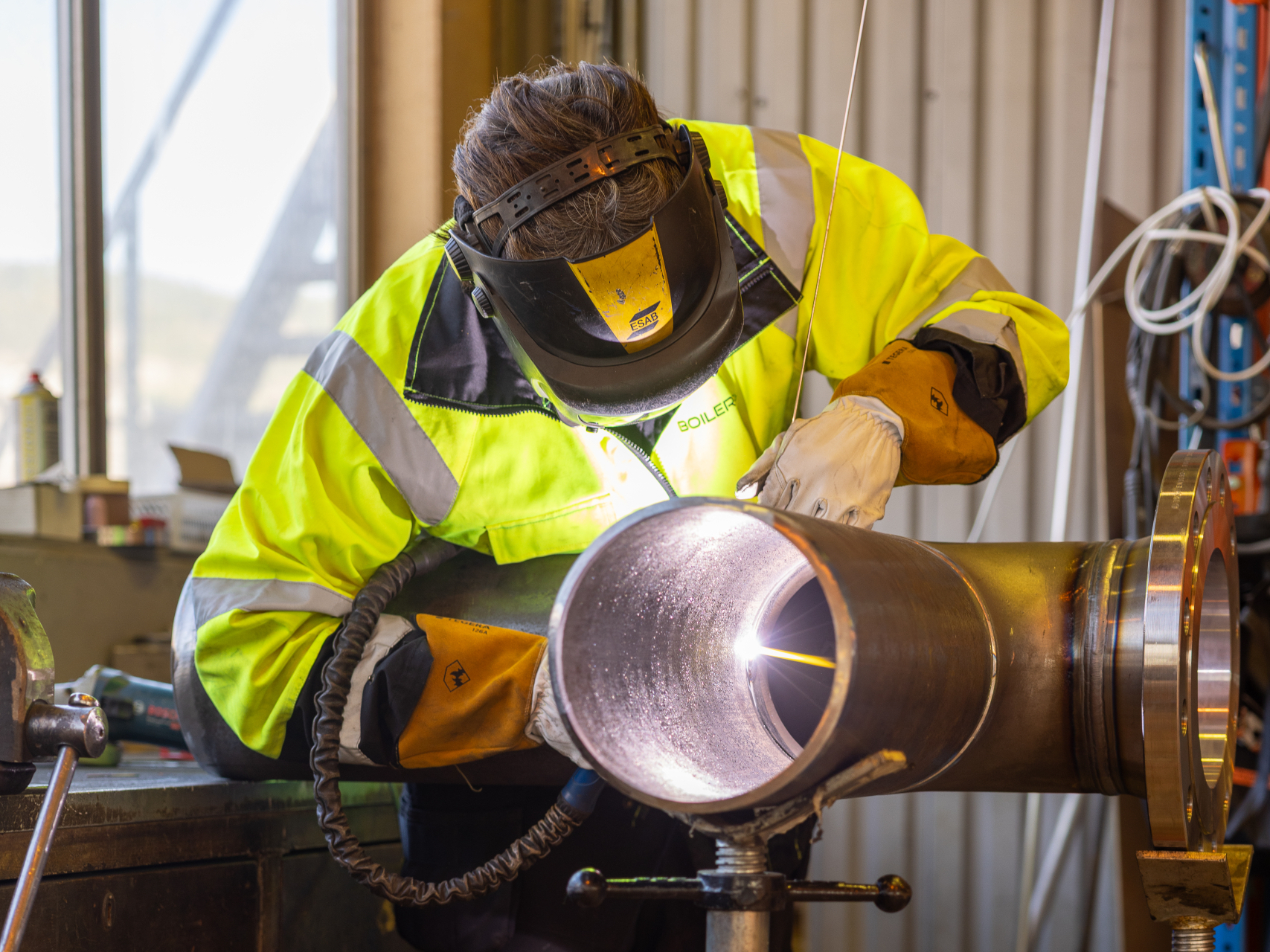 A welder wearing protective clothing and a welding helmet while welding an industrial pipe mounted on a pipe support in a workshop.